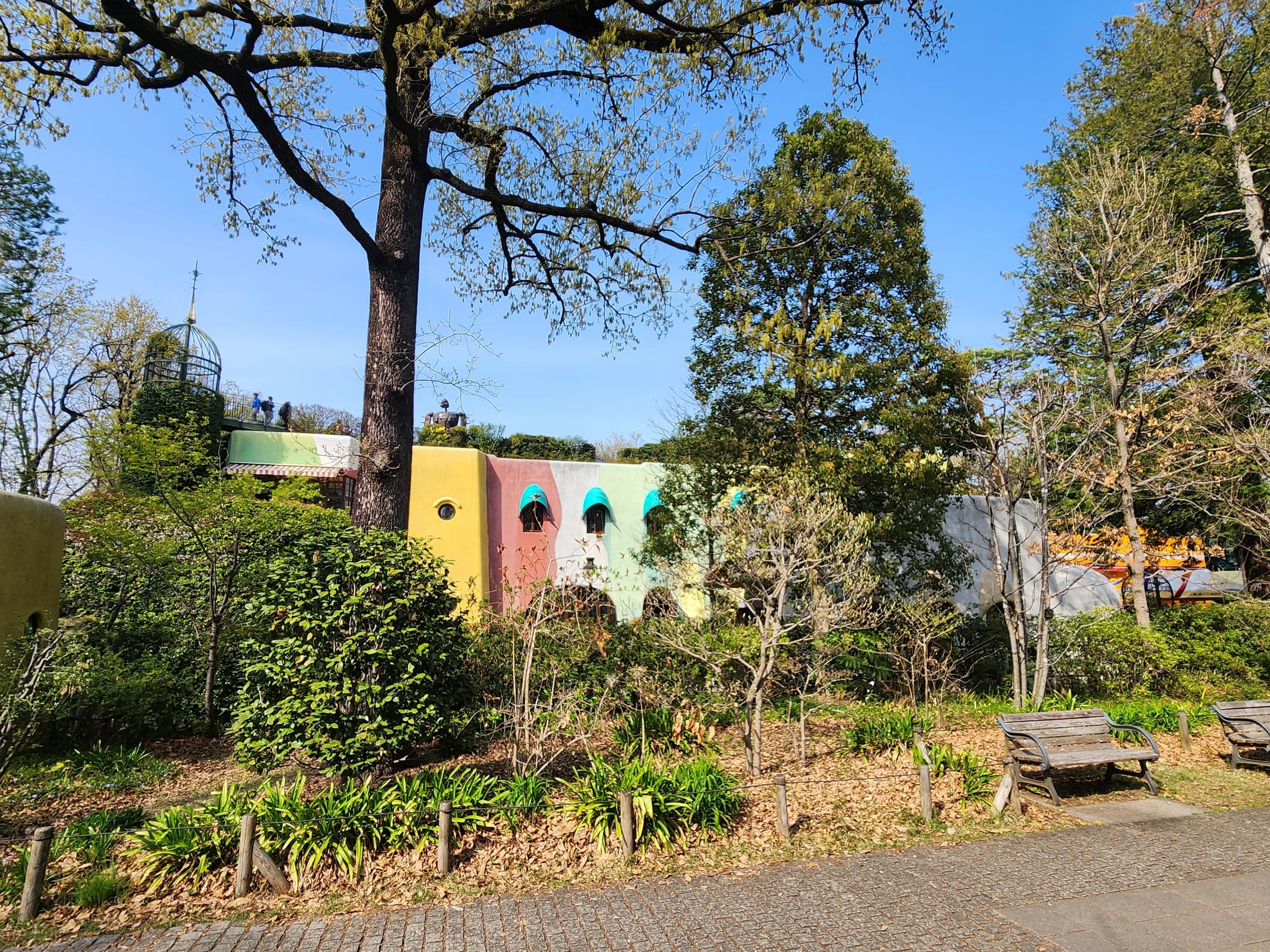 La fachada pastel del museo, amarilla, coral y menta, vista entre los árboles del parque Inokashira